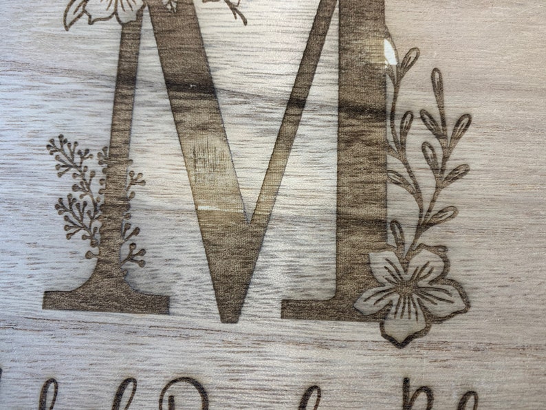 May include: A wooden sign with the letter "M" engraved in the center. The letter is surrounded by floral designs, including a flower and leaves.