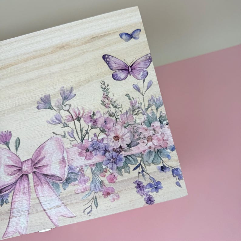 May include: Wooden box corner with a floral and butterfly design. The design features a large pink bow, purple butterflies, and a bouquet of pink, purple, and blue flowers. The wood has a light, natural finish.