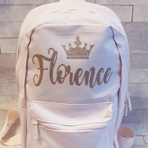 May include: A pink canvas backpack with a gold glitter crown and the name "Florence" written in gold glitter.