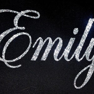 May include: The name "Emily" is spelled out in silver glitter on a black background.