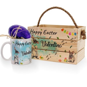 Puede incluir: Una caja de madera con asa, decorada con conejos, huevos de Pascua y el texto "Feliz Pascua Valentino". Una taza blanca con un diseño de conejo y el texto "Feliz Pascua Valentino" está delante de la caja.