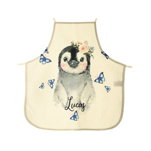 Puede incluir: Un delantal de cocina beige con un lindo pingüino de dibujos animados que lleva una corona de flores y está rodeado de mariposas azules. El delantal tiene el nombre "Lucas" escrito en cursiva debajo del pingüino.