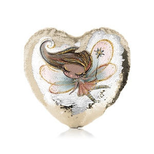 Könnte beinhalten: Ein herzförmiges Kissen mit einem umkehrbaren Pailletten-Design. Eine Seite hat eine goldene Pailletten-Oberfläche, während die andere Seite eine farbenfrohe Illustration einer Fee mit Flügeln und einem Zauberstab zeigt.
