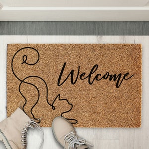 May include: A brown coir doormat with the word "Welcome" in black cursive lettering. A stylized black line drawing of a cat is on the left side of the mat.