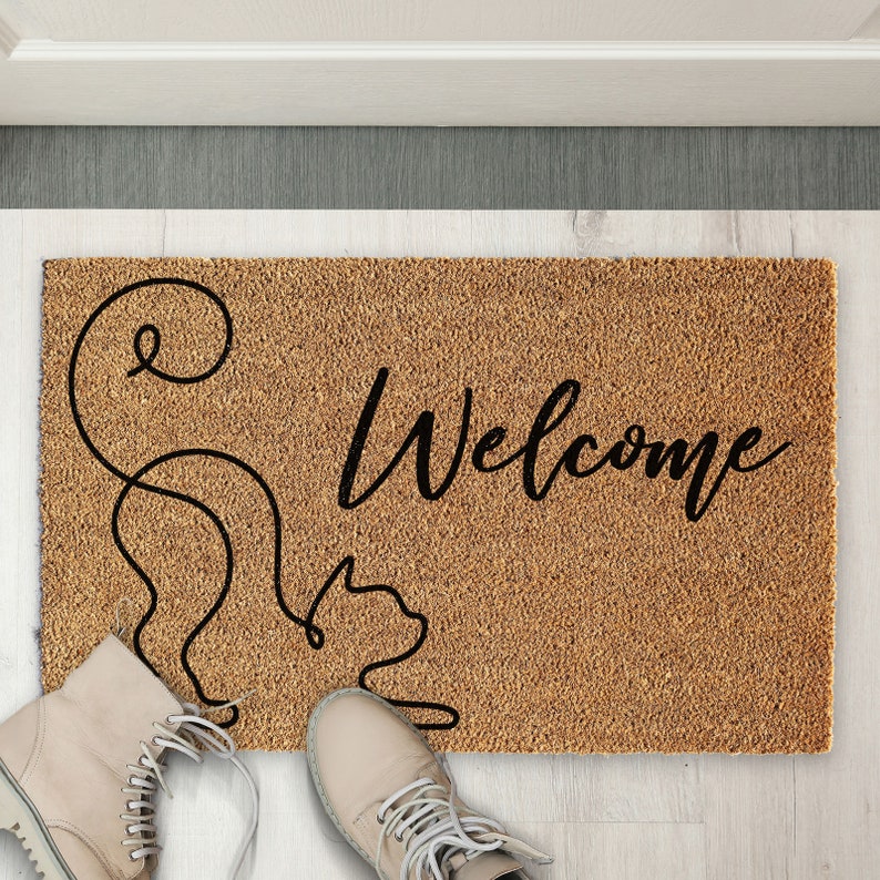May include: A brown coir doormat with the word "Welcome" in black cursive lettering. A stylized black line drawing of a cat is on the left side of the mat.