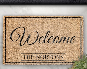 Puede incluir: Una alfombra de coco marrón con ribete negro y el texto "Welcome The Nortons" en una fuente cursiva.