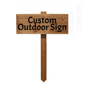May include: A rectangular wooden sign mounted on a vertical wooden stake. The sign features a natural wood grain texture and the black text Custom Outdoor Sign printed in a bold, sans-serif font across the center of the horizontal board.