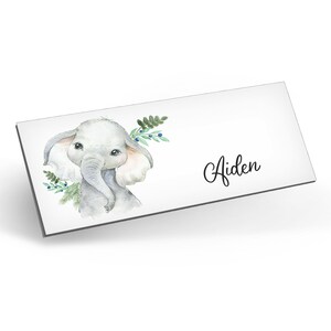 Puede incluir: Una ilustraci&oacute;n de acuarela de un lindo elefante gris con orejas rosadas y ojos azules. El elefante est&aacute; rodeado de hojas verdes y bayas azules. El nombre Aiden est&aacute; escrito en cursiva debajo del elefante.