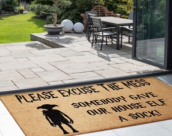 May include: A brown coir doormat with the text "Please excuse the mess, somebody gave our house elf a sock!" in black lettering. A black silhouette of a house elf is in the center of the mat.