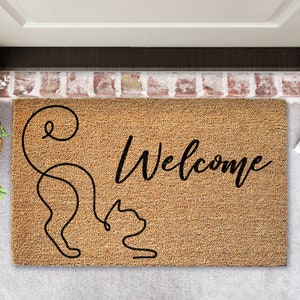 May include: A brown coir doormat with the word "Welcome" in black cursive lettering. A stylized black cat with a curled tail is on the left side of the mat.