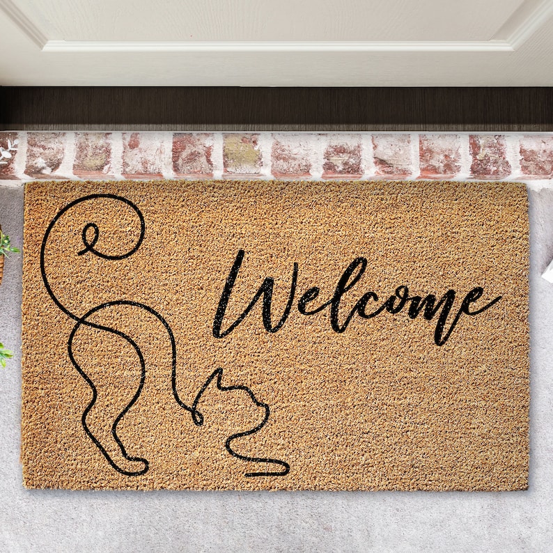 May include: A brown coir doormat with the word "Welcome" in black cursive lettering. A stylized black cat with a curled tail is on the left side of the mat.