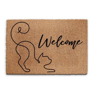 May include: A brown coir doormat with the word "Welcome" in black cursive lettering. A black line drawing of a cat with its tail curled around its body is on the left side of the mat.