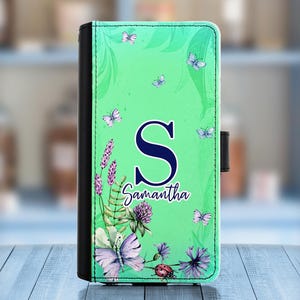 Puede incluir: Una funda de teléfono verde con un diseño floral y mariposas. La funda tiene una letra "S" azul y el nombre "Samantha" impreso en ella.