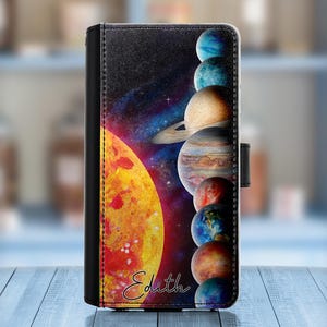 May include: A black leather phone case with a colorful galaxy design featuring planets and stars. The case has a flap that closes with a magnetic clasp. The name "Edith" is written in cursive on the case.