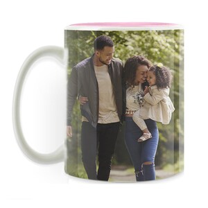 Può includere: Tazza in ceramica bianca con bordo interno rosa e manico curvo. La tazza presenta un'immagine avvolgente di una famiglia di tre persone, un uomo, una donna e un bambino, che camminano all'aperto in un parco. La famiglia sorride.