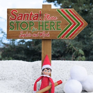 Op de afbeelding: Een houten bord met rode en groene pijlen die naar rechts wijzen. Op het bord staat "Santa! Please Stop Here for Sophia and Mark". Een kleine elfpop staat voor het bord en houdt een stok vast.