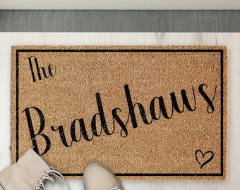 Puede incluir: Una alfombra de coco marrón con ribete negro y el texto "The Bradshaws" en letra cursiva. Un pequeño corazón se encuentra en la esquina inferior derecha de la alfombra.