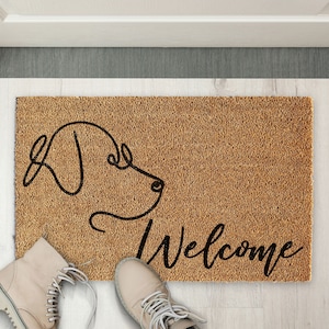 Puede incluir: Una alfombra de coco marrón con un dibujo de línea negra de la cabeza de un perro y la palabra "Bienvenido" en escritura negra.