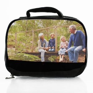 Puede incluir: Una bolsa de almuerzo negra con una impresión fotográfica rectangular en el panel frontal. La imagen muestra a una pareja mayor y dos niños sentados juntos en un puente de madera en un entorno forestal, rodeados de árboles y helechos.