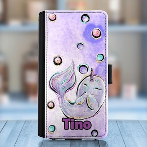May include: Purple phone case featuring a narwhal design with a horn and tail. The case has a watercolor background with bubbles and the name "Tino" in purple. The case is black on the spine and has a magnetic closure.