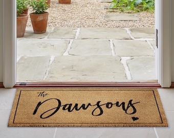 Puede incluir: Felpudo rectangular de fibra de coco natural. Presenta la frase "The Dawsons" en escritura negra, con un pequeño corazón. El felpudo está colocado en una puerta, con vistas a un jardín.