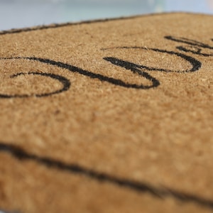 May include: A brown coir doormat with the word "Welcome" written in black cursive lettering.