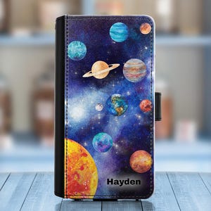 Op de afbeelding: Een gepersonaliseerde telefoonhoes met een aquarel-galactisch ontwerp met planeten en sterren. De hoes heeft de naam "Hayden" onderaan gedrukt.