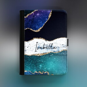 Peut inclure: Une housse de tablette en cuir noir avec un design inspir&eacute; de la galaxie, pr&eacute;sentant des couleurs bleues, violettes et turquoise avec des accents de paillettes dor&eacute;es. Le nom "Isabella" est &eacute;crit en paillettes dor&eacute;es sur le devant de l'&eacute;tui.