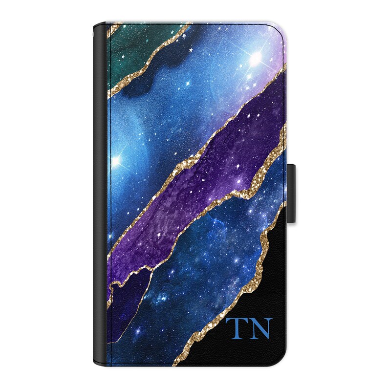 May include: A black phone case with a galaxy print and gold glitter accents. The case has a flap that closes with a snap. The initials "TN" are printed on the case.