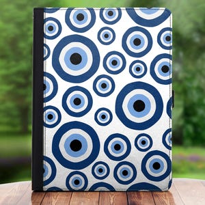 Puede incluir: Una funda para tableta con fondo blanco y un patrón de diseños de ojo turco azules y negros. La funda tiene una columna vertebral negra y un cierre magnético. El diseño se repite en toda la superficie.