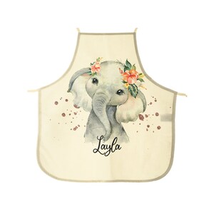 Puede incluir: Un delantal de cocina beige con una corona de flores en la cabeza de un elefante. El elefante es gris y tiene detalles rosas. El delantal tiene el nombre "Layla" impreso en él.