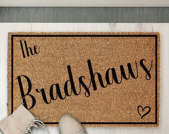 Puede incluir: Una alfombra de coco marrón con ribete negro y el texto "The Bradshaws" en letra cursiva. Un pequeño corazón se encuentra en la esquina inferior derecha de la alfombra.