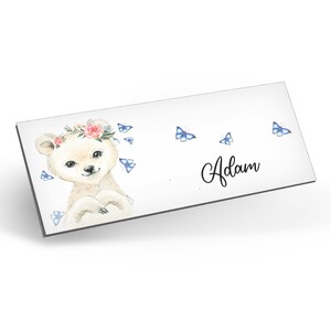 Puede incluir: Un oso blanco con una corona de flores rosas y mariposas azules, con el nombre "Adam" escrito en cursiva.