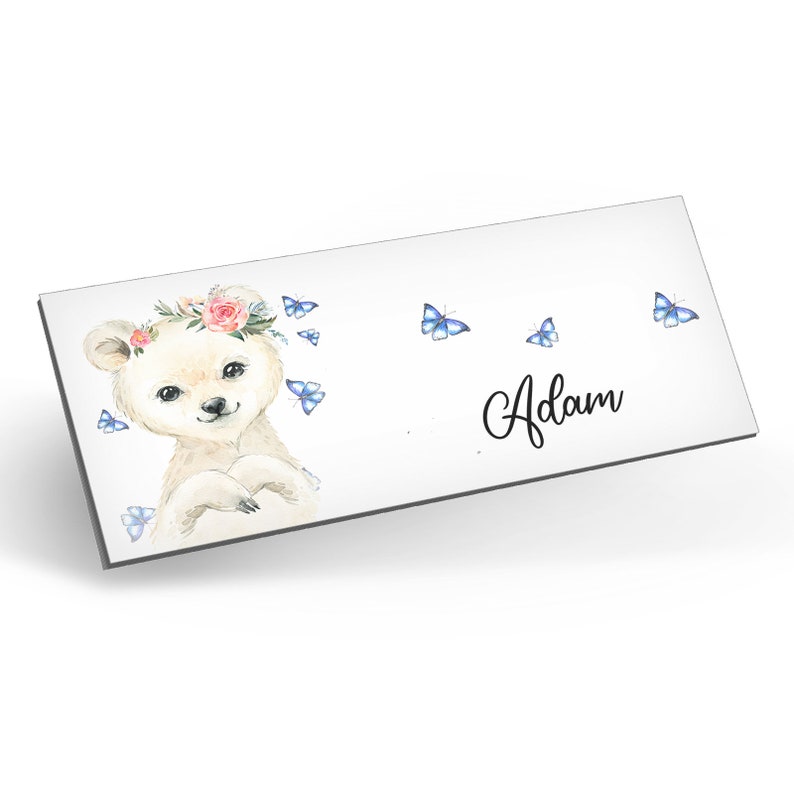 Puede incluir: Un oso blanco con una corona de flores rosas y mariposas azules, con el nombre "Adam" escrito en cursiva.