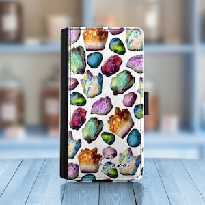 May include: A white phone case with a colorful pattern of gemstones. The case has a black leather flap with a snap closure. The name "Sara" is written in black cursive on the bottom of the case.
