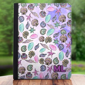 Puede incluir: Una funda para iPad de color morado y blanco con un patrón de conchas marinas y estrellas de mar. La funda tiene las iniciales "ML" en rosa en la esquina inferior derecha.