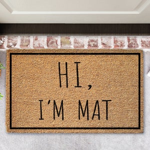 Puede incluir: Una alfombra de coco marrón con ribete negro y el texto "HI, I'M MAT" en negro.