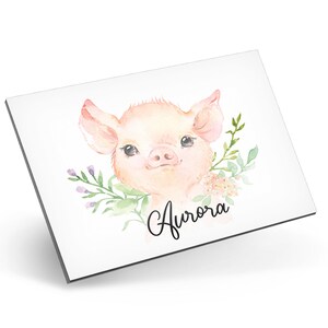 Puede incluir: Una ilustraci&oacute;n de acuarela de un lech&oacute;n rosado con una corona de flores alrededor de su cabeza. El lech&oacute;n est&aacute; mirando al espectador con una expresi&oacute;n dulce. El nombre "Aurora" est&aacute; escrito en cursiva debajo del lech&oacute;n.