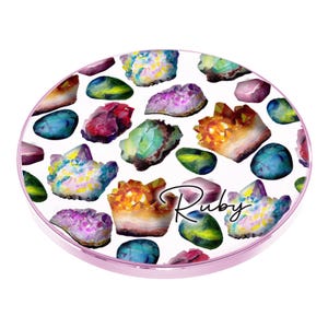May include: A round, white phone case with a pink rim. The case is decorated with a pattern of colorful gemstones, including amethyst, citrine, and turquoise. The word "Ruby" is written in cursive on the case.