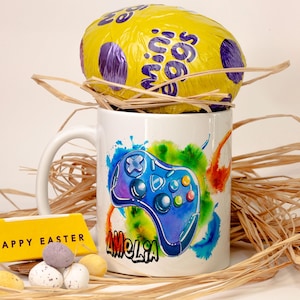 Könnte beinhalten: Eine weiße Tasse mit einem blau-violetten Videospiel-Controller-Design und dem Namen "Amelia". Ein großes gelbes Osterei liegt auf der Tasse, umgeben von Stroh. Ein gelbes Schild liest "Frohe Ostern". Kleine Ostereier sind ebenfalls vorhanden.