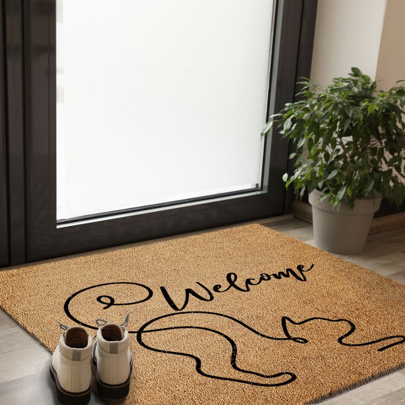 May include: A brown coir doormat with the word "Welcome" written in black cursive script. A black outline of a cat with its tail curled into a heart shape is on the doormat.