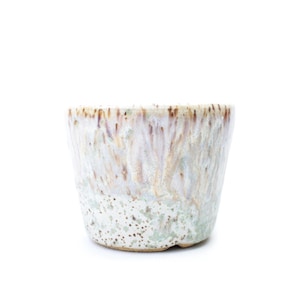 May include: A small ceramic cup with a distinctive glaze. The cup has a white base with a speckled pattern and a blend of brown, green, and purple hues that drip down the sides. The rim has a textured appearance.