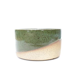 May include: A cylindrical ceramic bowl featuring a two-tone design with a speckled moss green glaze on the upper portion and a speckled cream-colored base. The exterior surface has a visible texture with dark brown flecks throughout the finish.