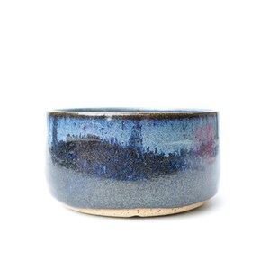 May include: A round ceramic bowl featuring a reactive glaze with deep blue, navy, and charcoal gray tones. The exterior surface displays a mottled, speckled texture with a smooth finish, while the base remains unglazed with a light tan color.