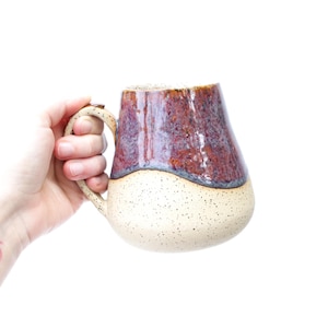 May include: A ceramic mug with a speckled white glaze and a brown and purple glaze on the top half. The mug has a rounded shape and a handle.