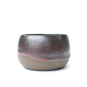 Könnte beinhalten: Eine kleine, runde Keramikschale mit zweifarbiger Oberfläche. Der obere Teil ist mit einer metallisch schimmernden Glasur in tiefen Rot- und Dunkelbrauntönen versehen, während der untere Teil eine matte, gesprenkelte, hellbraune Tontextur zeigt.