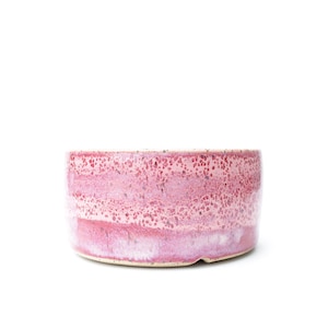 Könnte beinhalten: Eine zylindrische Keramikschale mit einer gesprenkelten Glasur in verschiedenen Rosa- und Weißtönen. Die Außenfläche zeigt ein strukturiertes, geflecktes Muster mit dunkleren Magentapunkten auf einem helleren, zartrosa Untergrund.