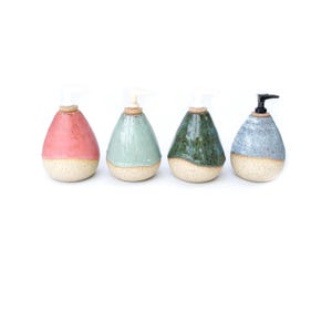 May include: Four ceramic soap dispensers in a row, each with a unique color and design. The dispensers are teardrop-shaped, with a speckled beige base and a colored top. Colors include pink, seafoam green, dark green, and blue with a black pump.