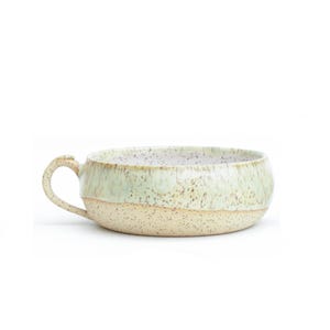 May include: A wide, low-profile ceramic bowl with a single handle, featuring a speckled cream-coloured base and a smooth, pale mint green glazed upper section with darker brown speckles throughout the entire exterior and interior surface.