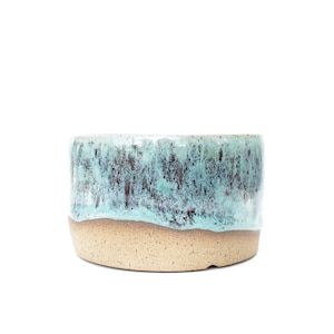 May include: A cylindrical ceramic bowl featuring a two-tone glaze. The upper portion displays a mottled turquoise and dark brown drip pattern, while the lower section consists of a speckled, unglazed beige clay base with a smooth texture.